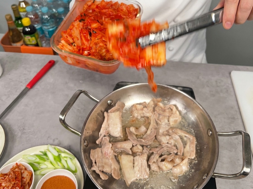 Leuchtend rotes Kimchi wird zu den angebratenen Schweinebauchscheiben in die Pfanne gegeben.
