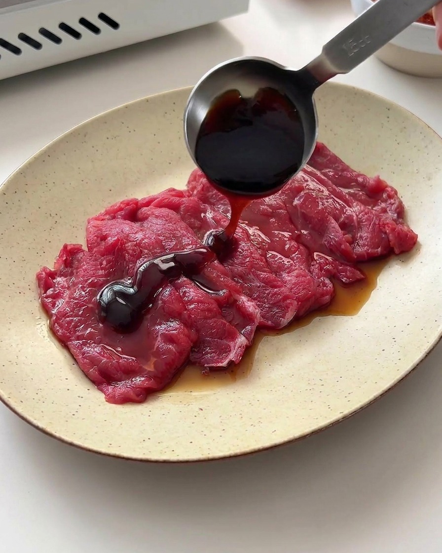 Sojasauce und Austernsauce werden aus einem Messlöffel über das rohe Rindfleisch auf dem Teller gegossen.