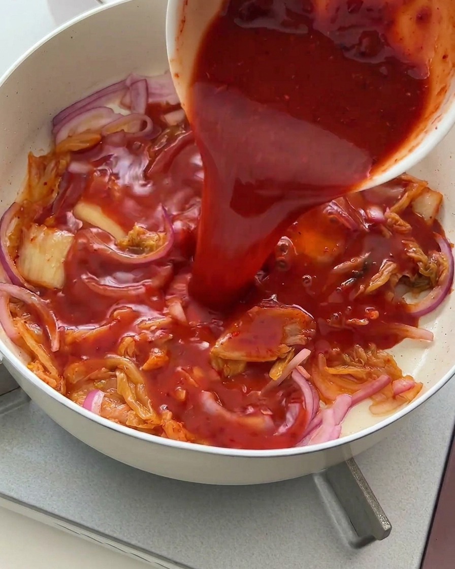 Eine kräftige rote Würzsauce wird aus einer Schüssel über das Kimchi und die Zwiebeln gegossen.