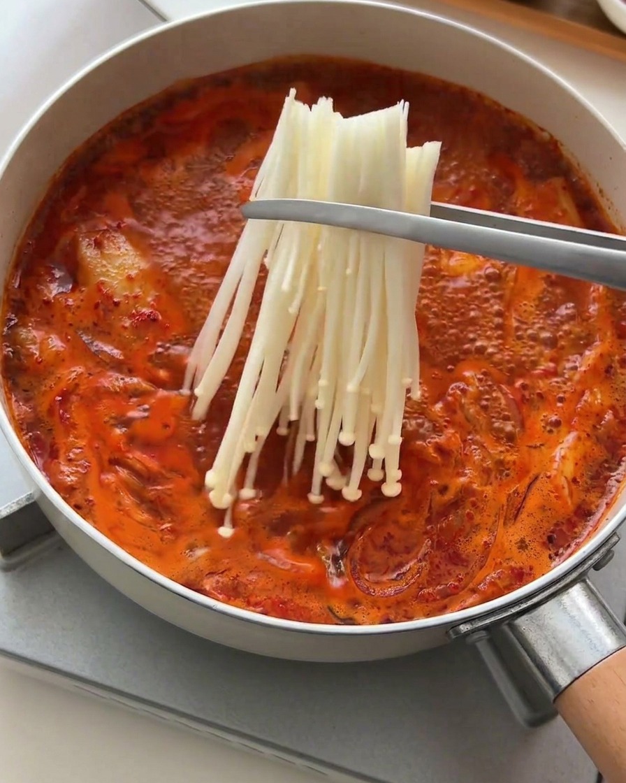 Eine Metallzange setzt ein Bündel weißer Enoki-Pilze in die sprudelnde rote Suppe.