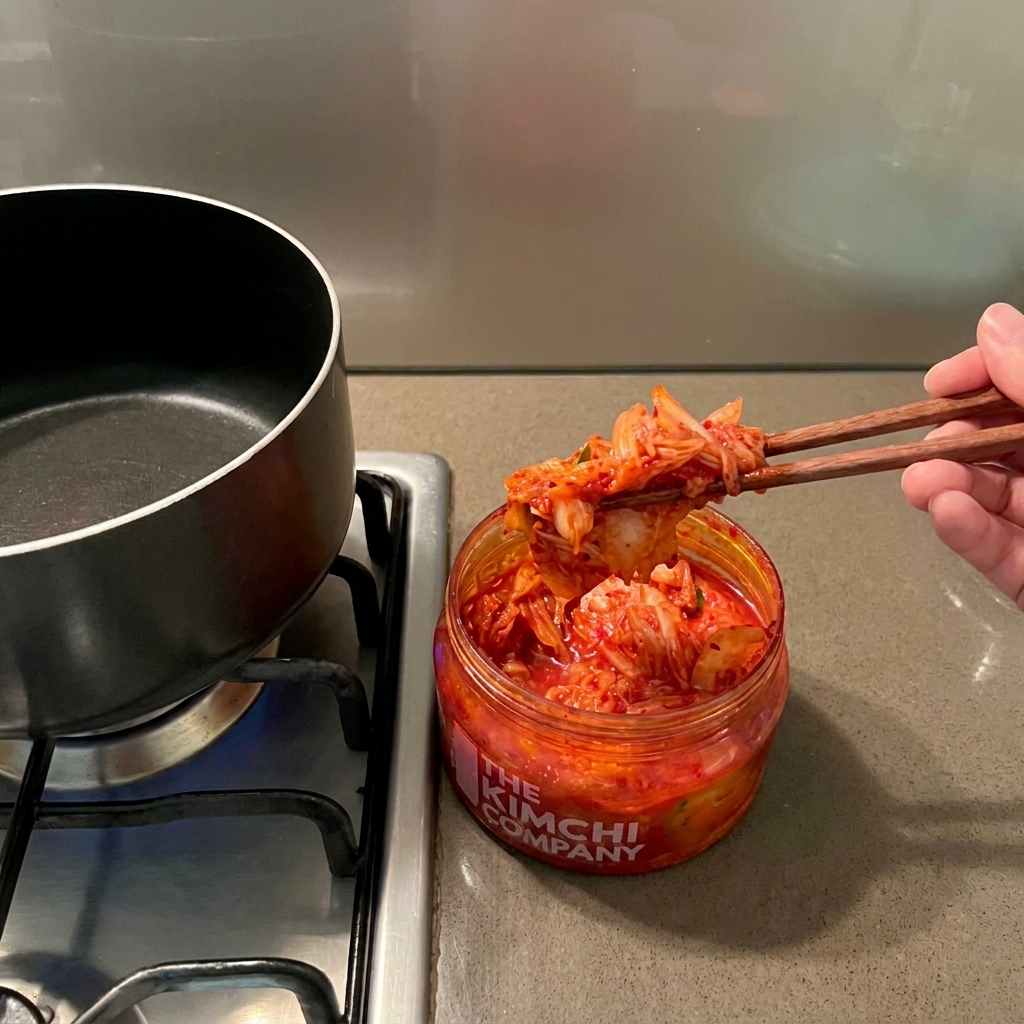 Eine Hand, die mit Essstäbchen rotes Kimchi aus einem Glas in einen schwarzen Kochtopf füllt.