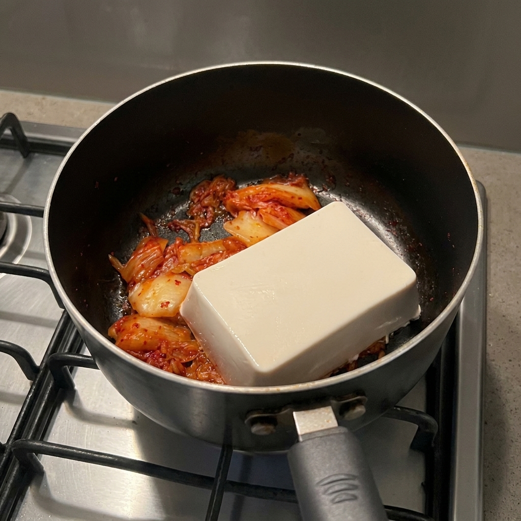 Ein ganzer weißer Tofublock liegt auf angebratenem roten Kimchi in einem Kochtopf.