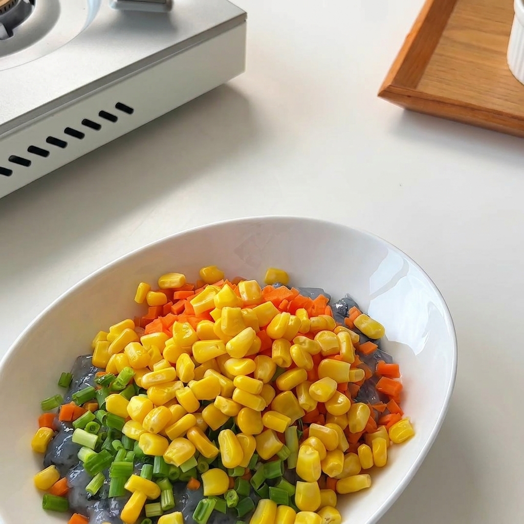 Eine kleine Schüssel mit goldenen Maiskörnern wird in eine Schüssel mit Garnelenpaste, Karotten und Zwiebeln geleert.