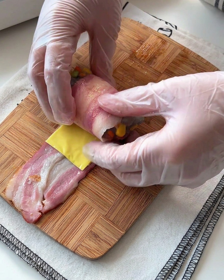 Hände rollen vorsichtig einen Speckstreifen um die Garnelen-Käse-Füllung auf einer Holzoberfläche.