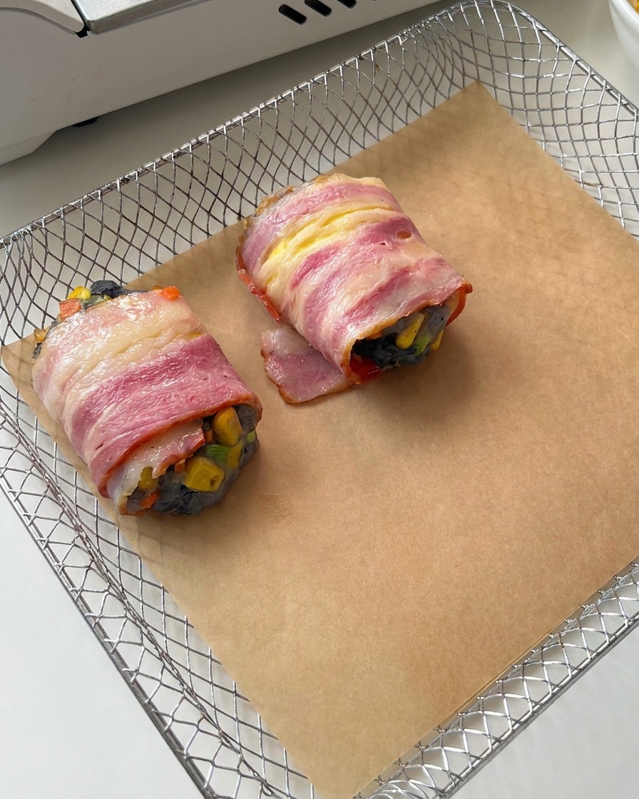Zwei rohe Speck-Garnelen-Rollen liegen auf Backpapier in einem Korb der Heißluftfritteuse.