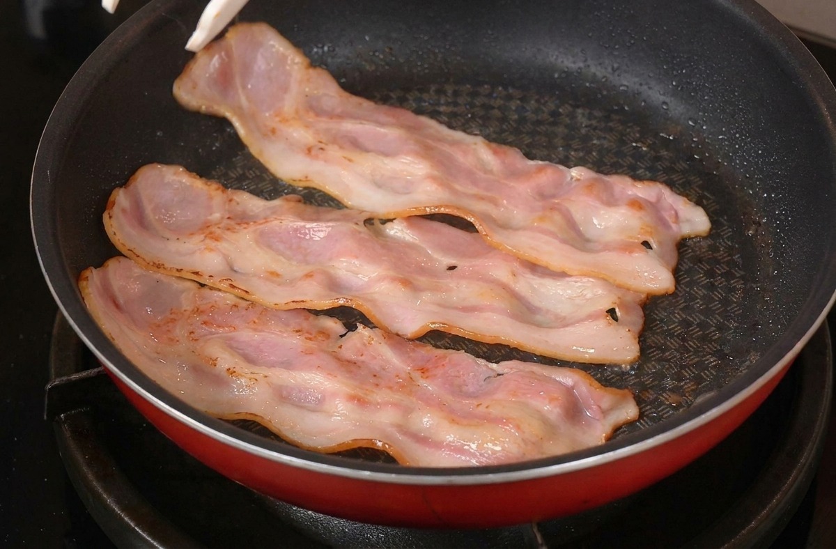 Eine Grillzange wendet angebratene, sich wellende Speckstreifen in einer roten Pfanne auf dem Herd.