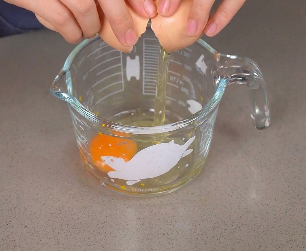 Hände schlagen ein braunes Ei über einem Glas auf, in dem sich bereits ein rohes Ei befindet.