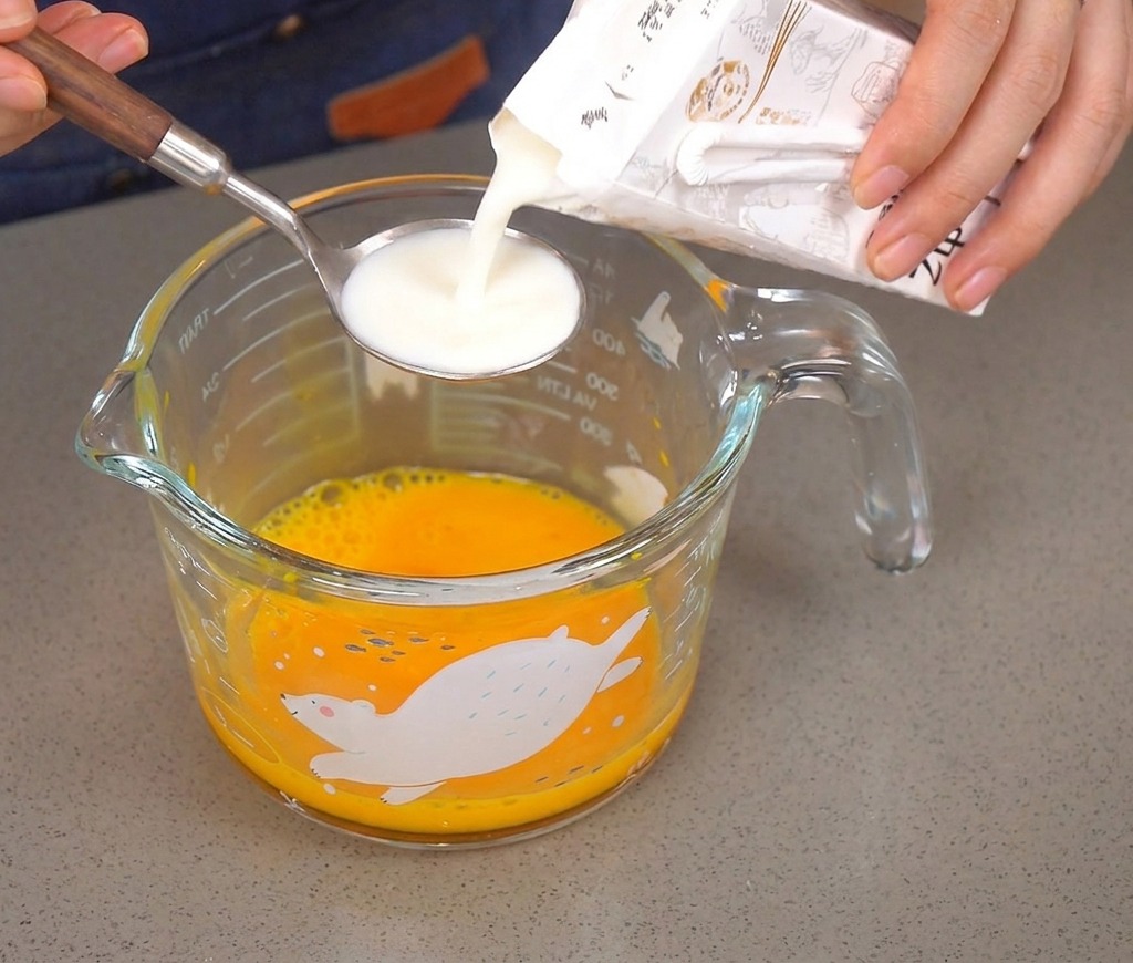 Milch wird aus einem Karton in einen silbernen Löffel über einem Becher mit verquirlten Eiern gegossen.