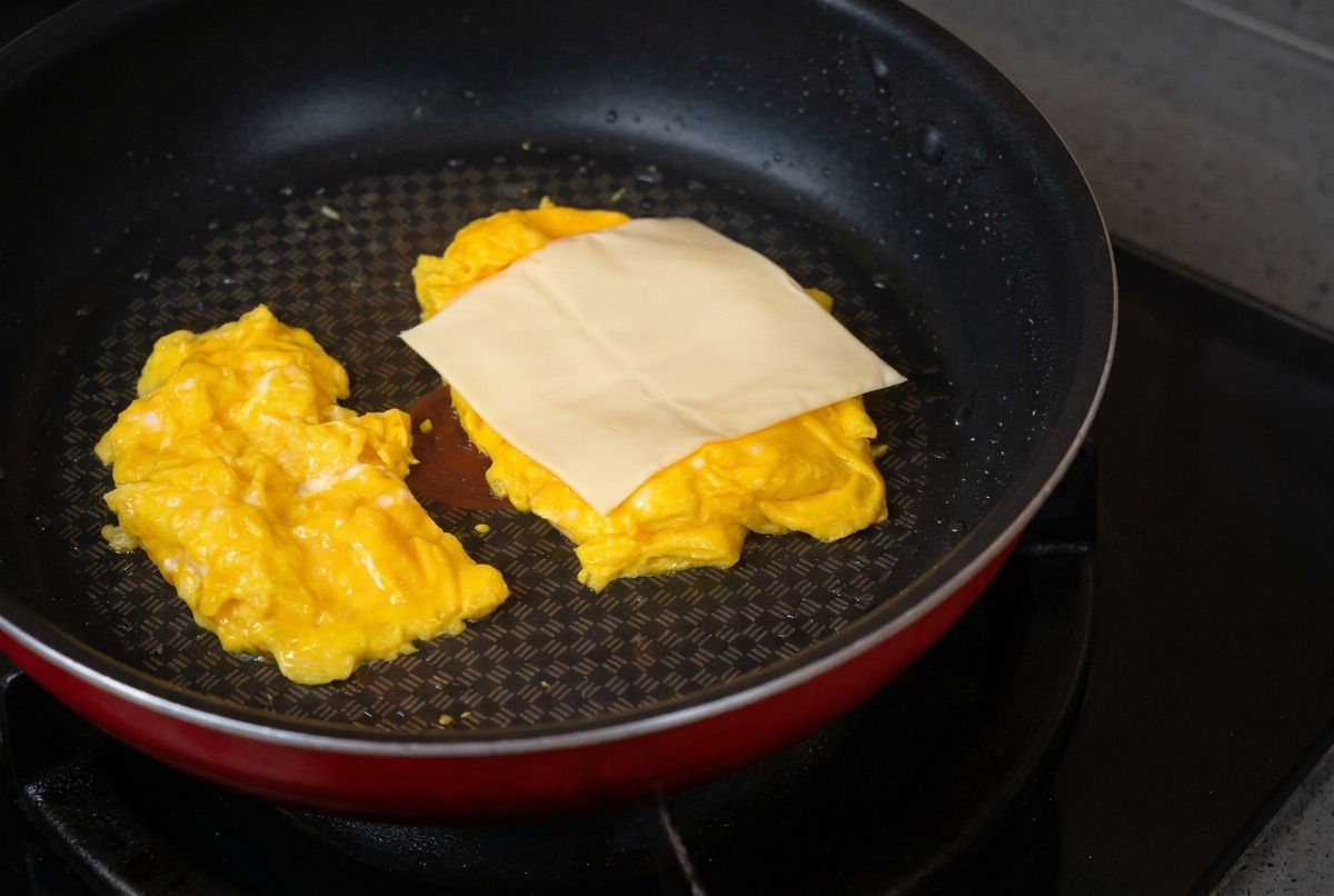 Eine beschichtete Pfanne mit zwei Portionen Rührei, auf die gerade eine quadratische Scheibe Käse gelegt wird.
