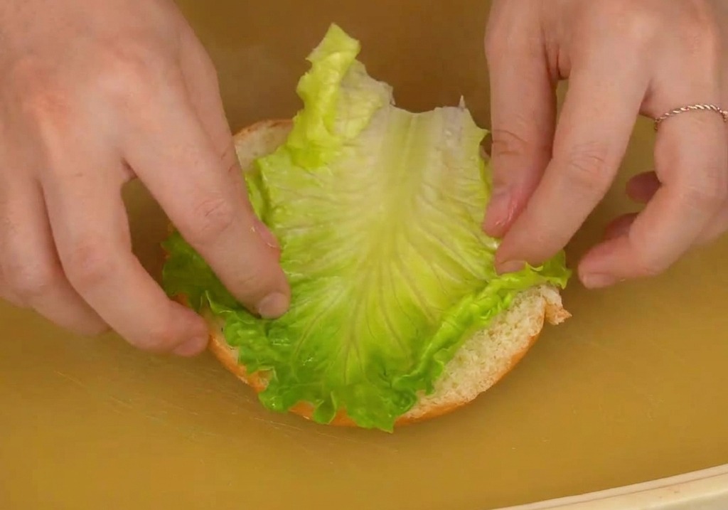 Zwei Hände legen vorsichtig ein großes, frisches grünes Salatblatt auf die untere Hälfte eines Burgerbrötchens.