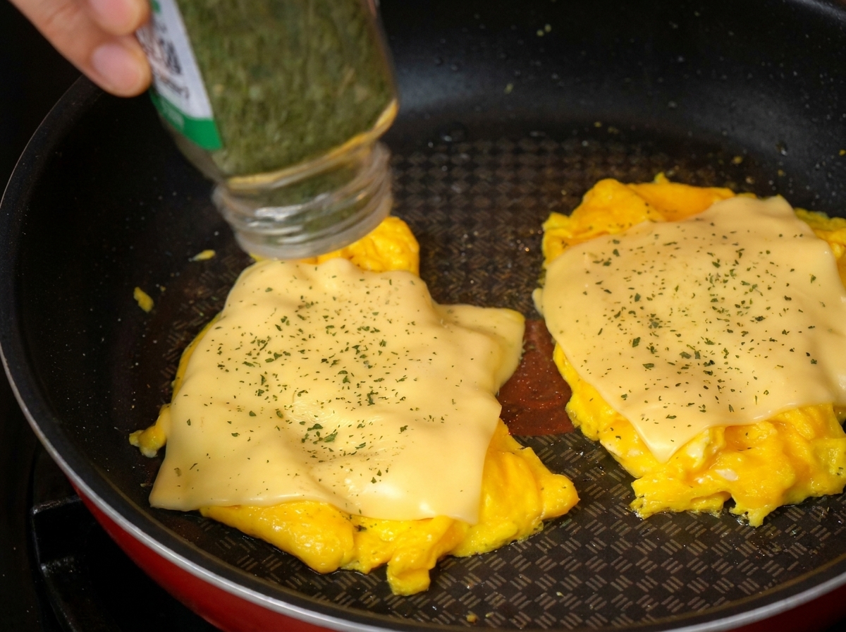 Nahaufnahme von getrockneter Petersilie, die aus einem Gewürzstreuer auf das Rührei mit Käse gestreut wird.