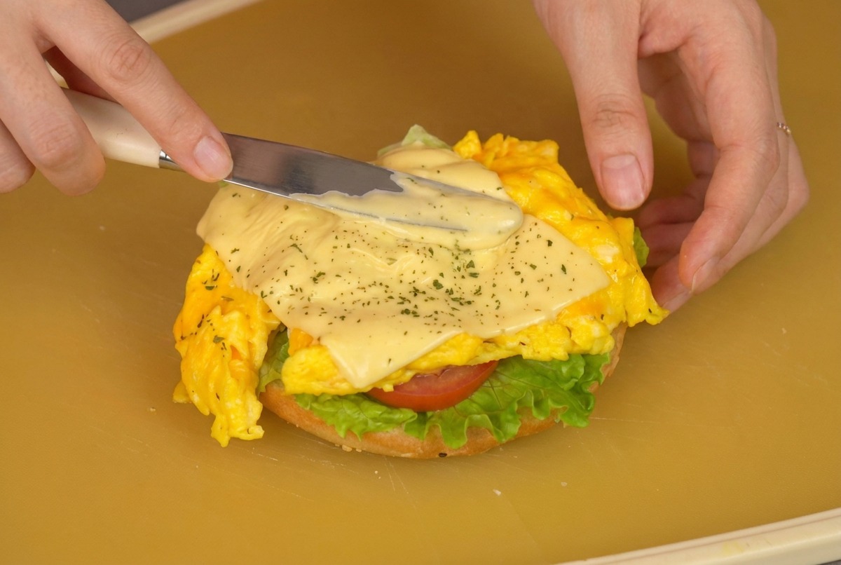 Nahaufnahme einer Person, die mit einem Pfannenwender eine Portion Rührei mit Käse auf das mit Salat und Tomate belegte Brötchen hebt.