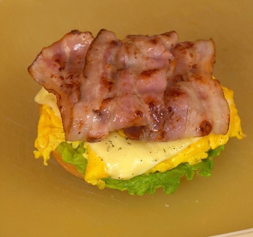 Ein fertig geschichteter Burger auf einem Schneidebrett mit Salat, Tomate, Käse-Rührei und knusprigem Speck.