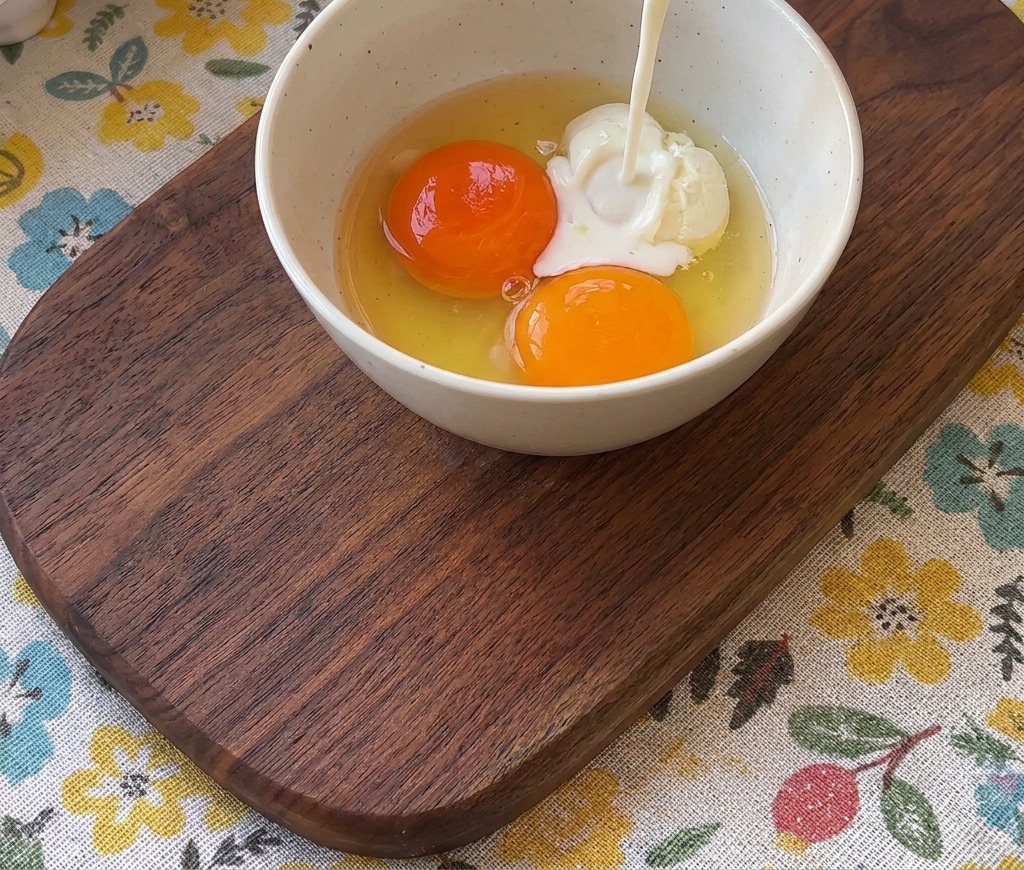 Milch wird aus einem Glas in eine Schüssel mit zwei leuchtend orangefarbenen Eigelben gegossen.