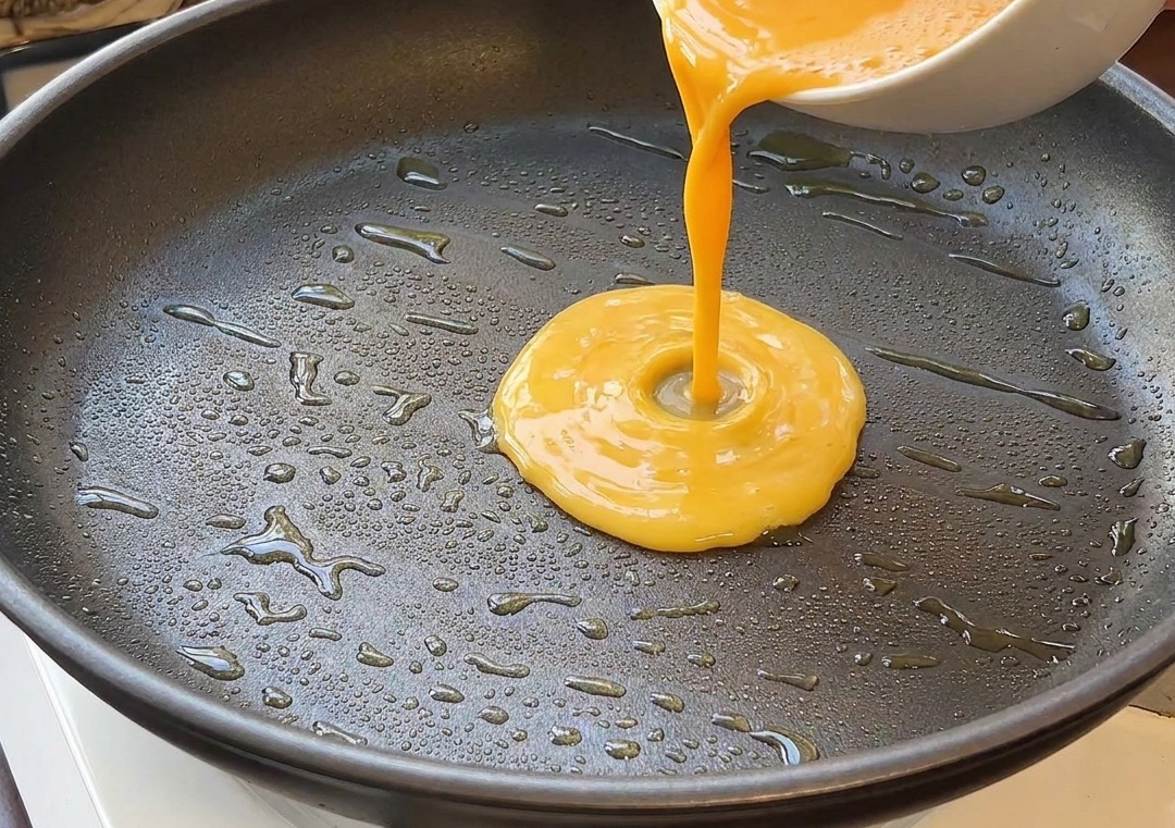 Gelbe Eiermasse wird aus einer weißen Schüssel in eine heiße Pfanne mit glänzendem Öl gegossen.
