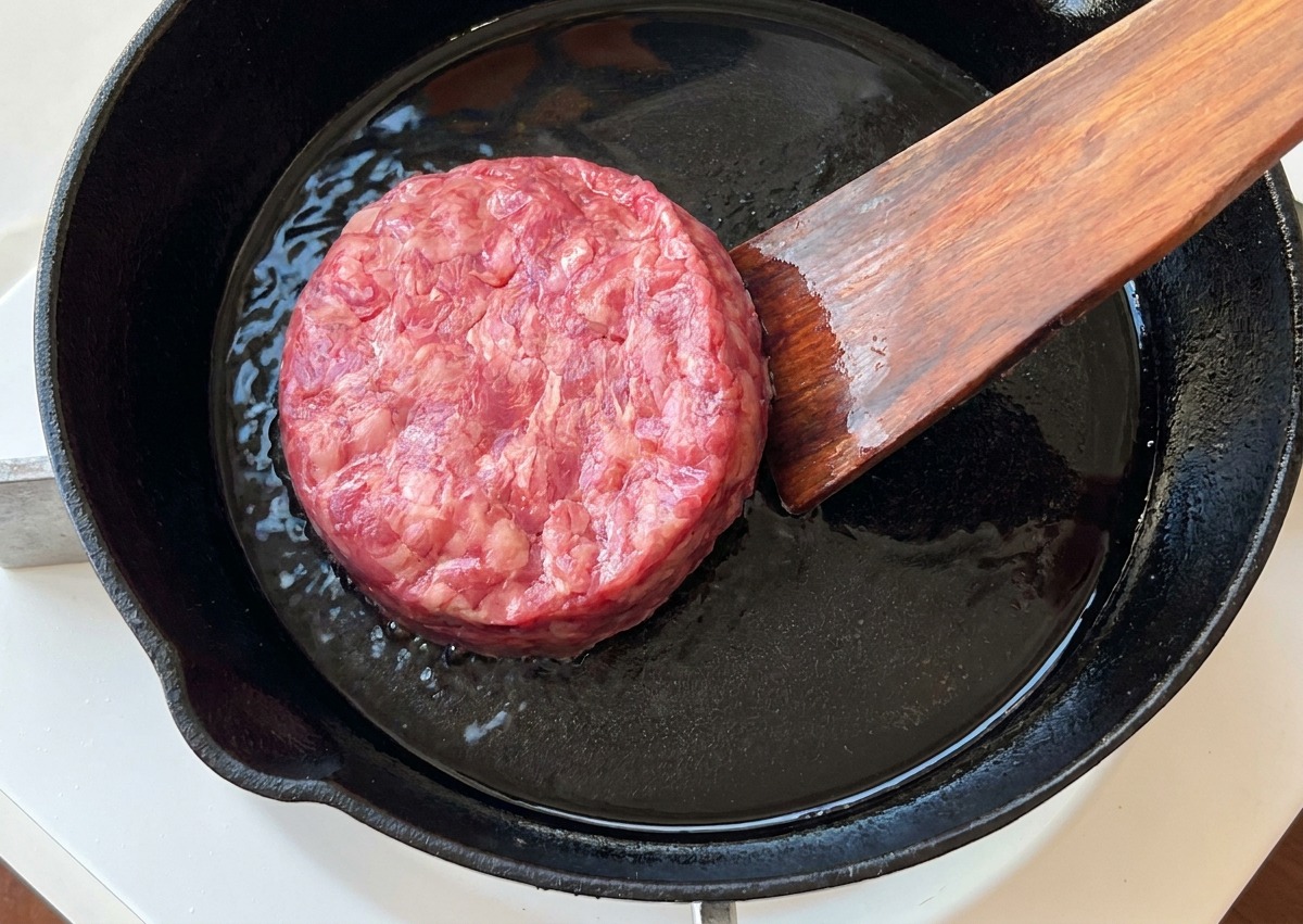 Ein rohes Rinderhack-Patty wird mit einem Pfannenwender in eine vorgeheizte gusseiserne Pfanne gelegt.