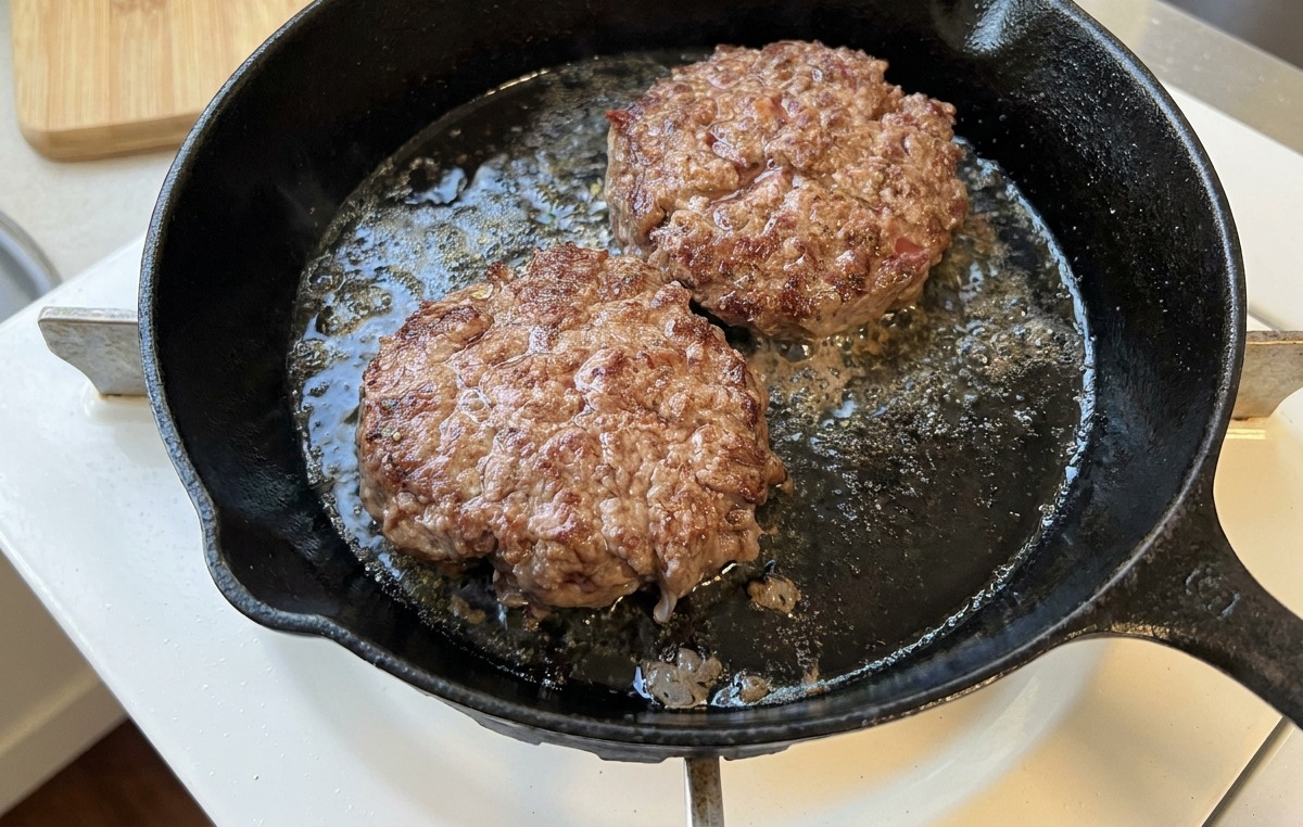 Zwei brutzelnde Fleisch-Patties in einer gusseisernen Pfanne mit einer tiefbraunen, karamellisierten Kruste.