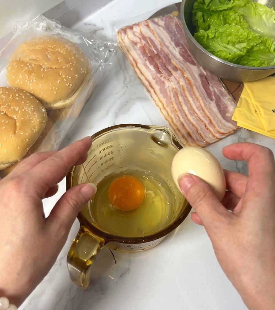 Eine Person schlägt ein rohes Ei in einen Messbecher auf, in dem sich bereits ein Ei befindet; im Hintergrund sind die Burgerzutaten zu sehen.