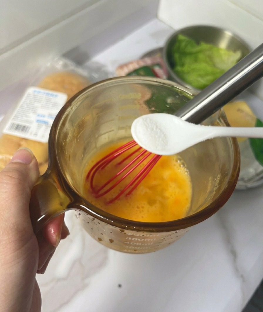 Eine Hand hält einen kleinen weißen Löffel mit Salz über einen Becher mit verquirlten gelben Eiern und einem roten Schneebesen.