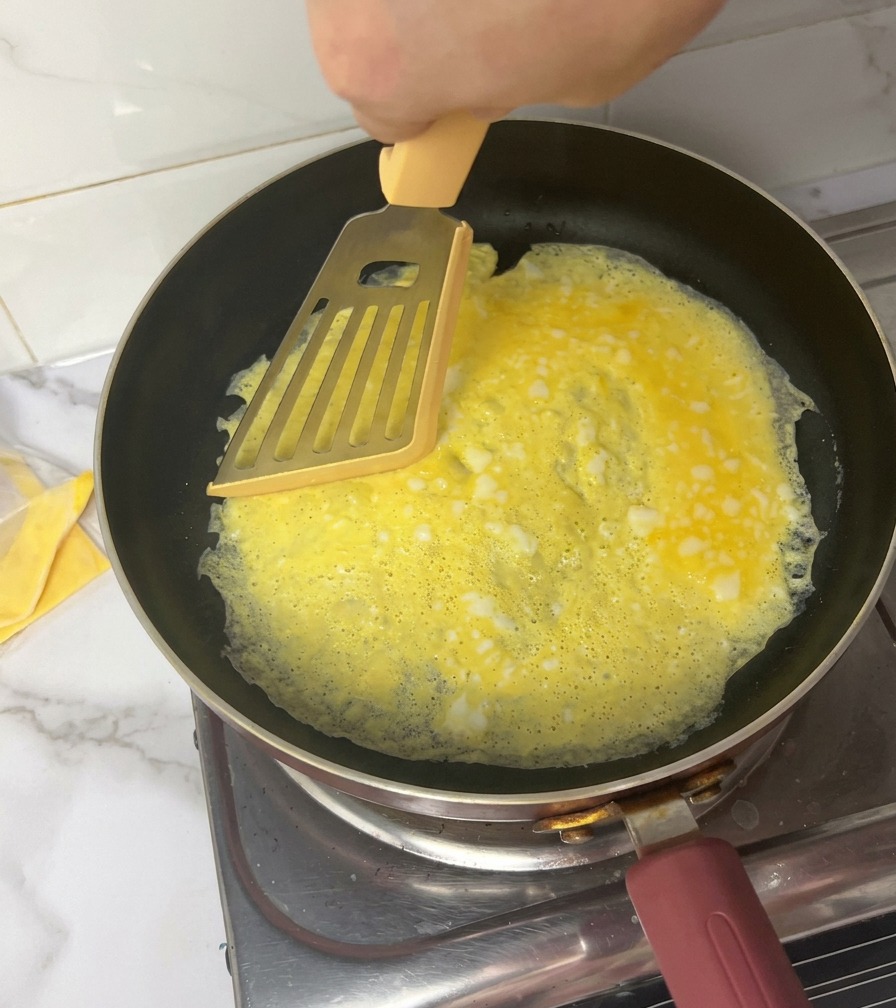 Eine Hand teilt ein dünnes gelbes Omelett in einer schwarzen Pfanne mit einem gelben Pfannenwender in vier Teile.