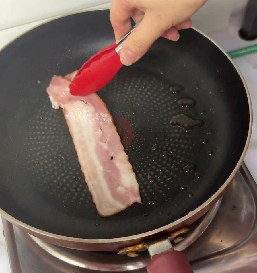 Eine Hand legt mit einer Grillzange einen langen Streifen rohen Speck in eine schwarze, beschichtete Pfanne.