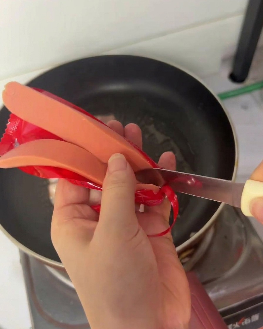 Eine Person schneidet ein Hot-Dog-Würstchen mit einem kleinen Messer längs über einer Pfanne durch.