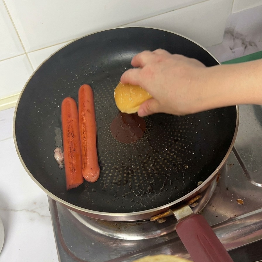 Eine Person legt die Oberseite eines Sesam-Burgerbrötchens mit der Schnittfläche nach unten neben zwei bräunende Würstchen in eine Pfanne.
