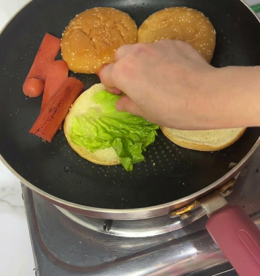 Eine Hand legt ein hellgrünes Salatblatt auf die geröstete Seite eines Burgerbrötchens in der Pfanne.