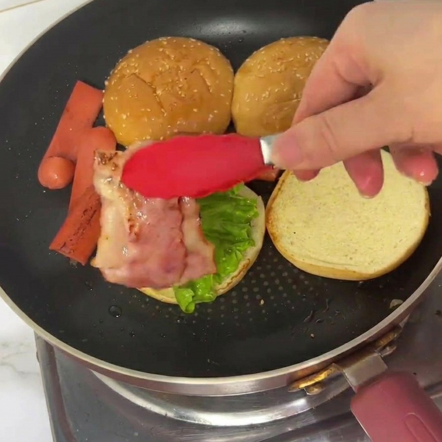 Eine rote Zange legt einen Streifen gebratenen Speck auf ein Burgerbrötchen, das bereits mit Salat belegt ist.