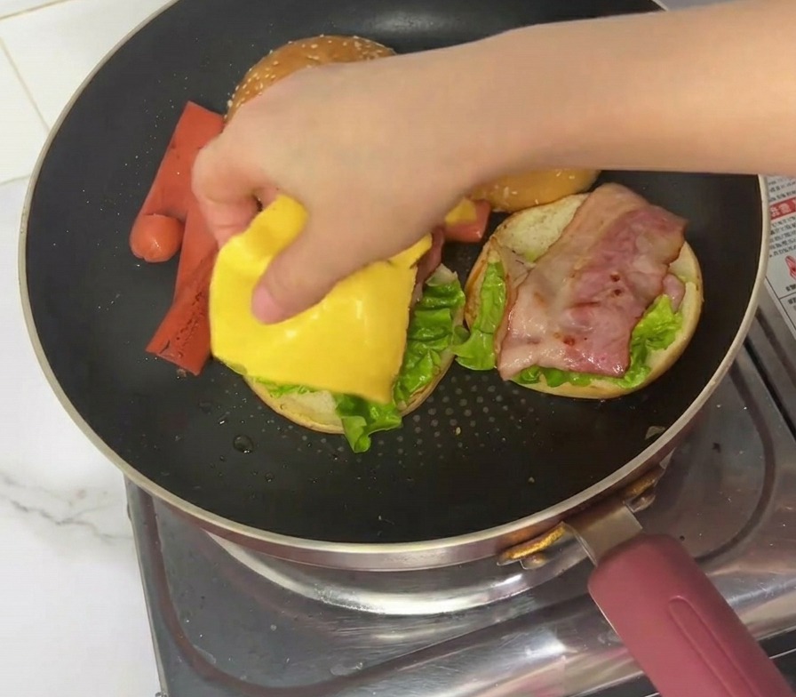 Eine Hand legt eine Scheibe gelben Schmelzkäse auf einen Burger-Zusammenbau in der Pfanne.