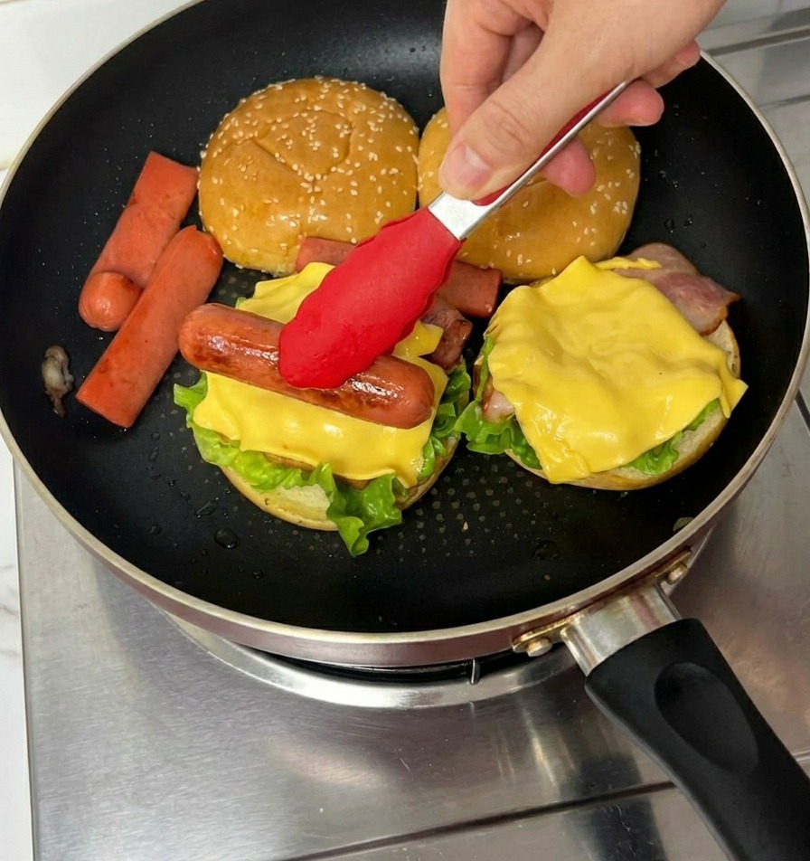 Eine rote Zange legt ein gebratenes Hot-Dog-Würstchen auf eine Käsescheibe im Burger.