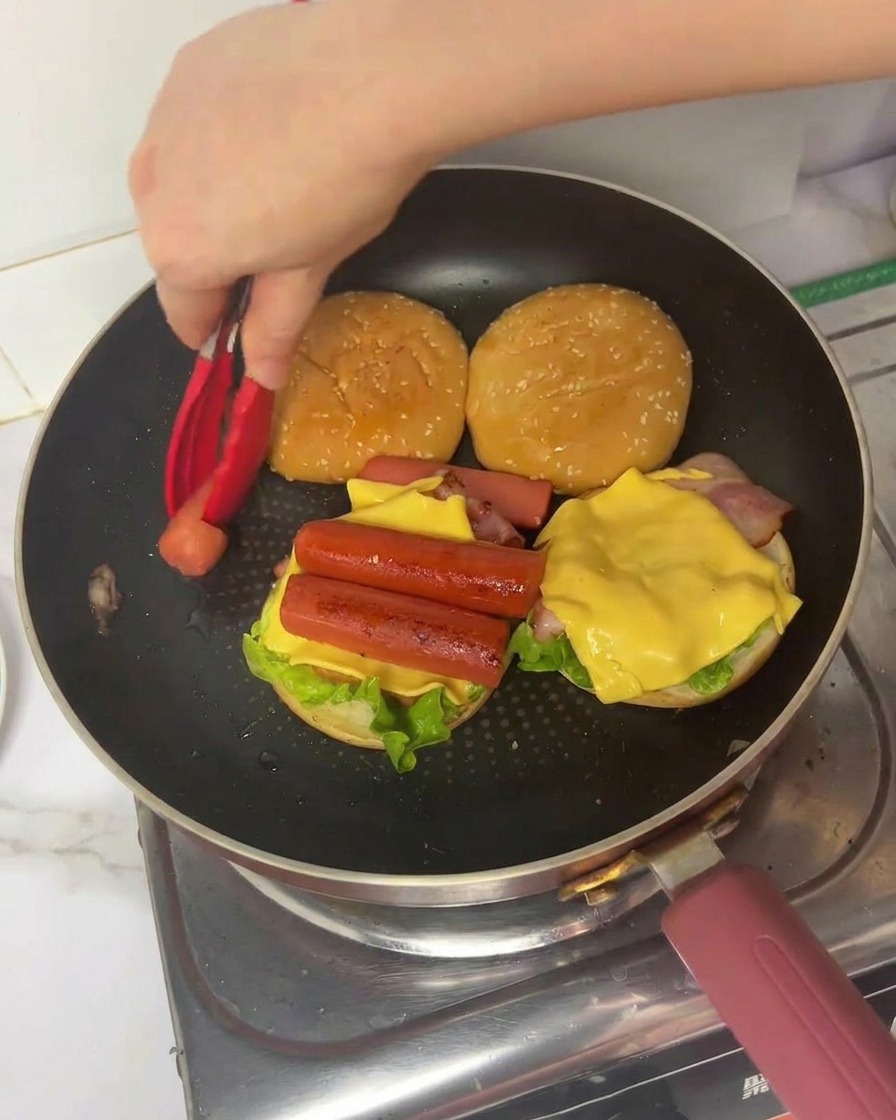 Eine Hand legt mit einer roten Zange ein Stück gefaltetes Ei auf einen geschichteten Burger in der Pfanne.