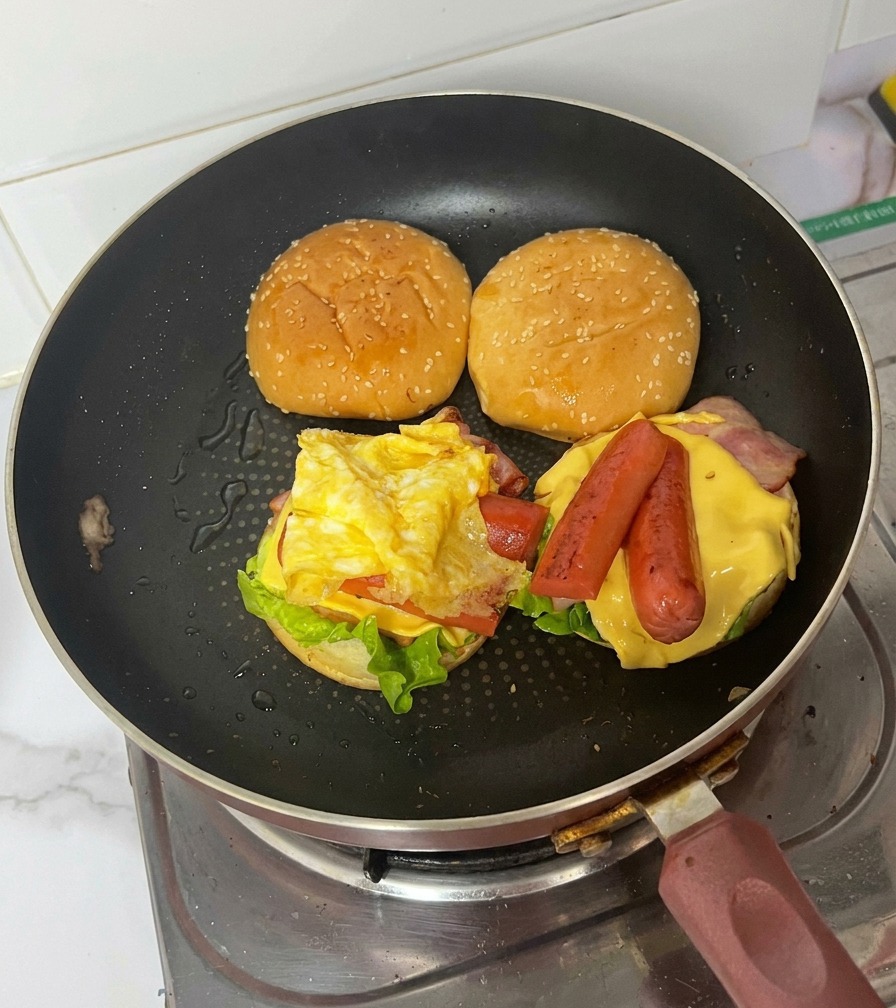 Ein fertiger Burger in der Pfanne zeigt das gefaltete Ei über Würstchen, Käse, Speck und Salat.