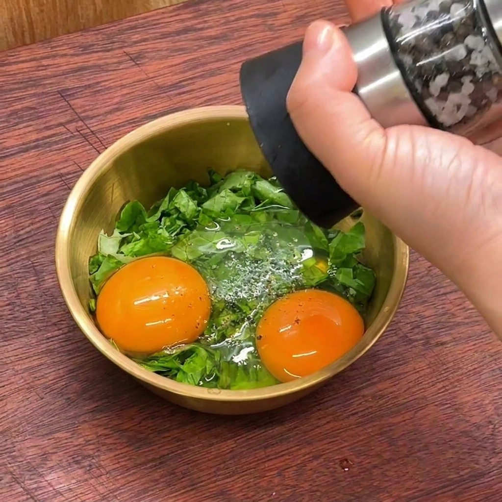 Eine Hand benutzt eine Pfeffermühle, um zwei rohe Eigelbe und geschnittenen Salat in einer Schüssel zu würzen.