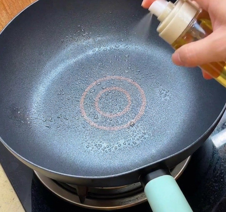 Eine Person besprüht eine schwarze beschichtete Bratpfanne mit Öl aus einer weißen Sprühflasche.