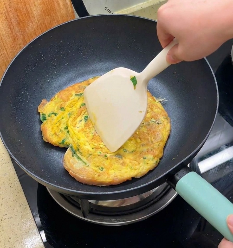 Ein Pfannenwender wendet ein goldbraunes Omelett samt Burgerbrötchen in einer Pfanne.