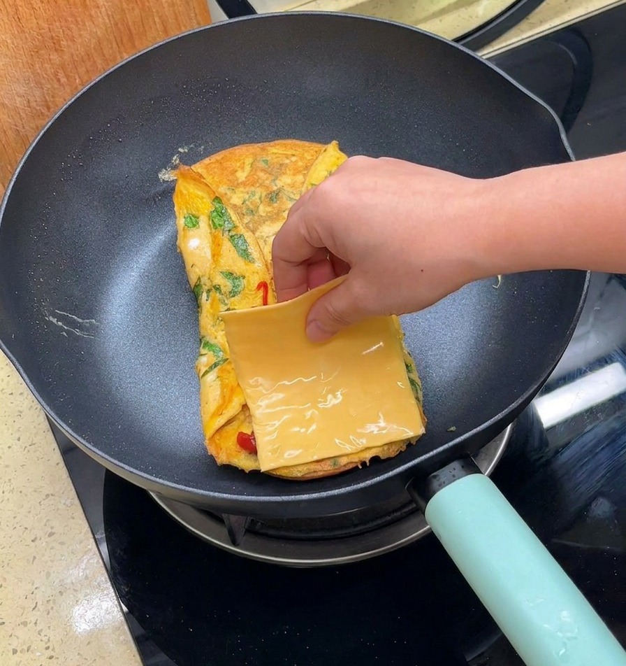 Eine Hand legt eine quadratische Scheibe gelben Käse auf die gefaltete Eischicht in der Pfanne.