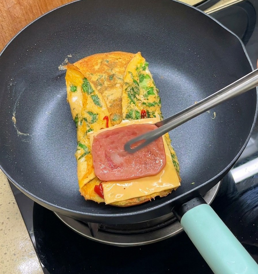 Eine Metallzange legt eine Scheibe gegarten Speck auf den Käse über dem Omelett.