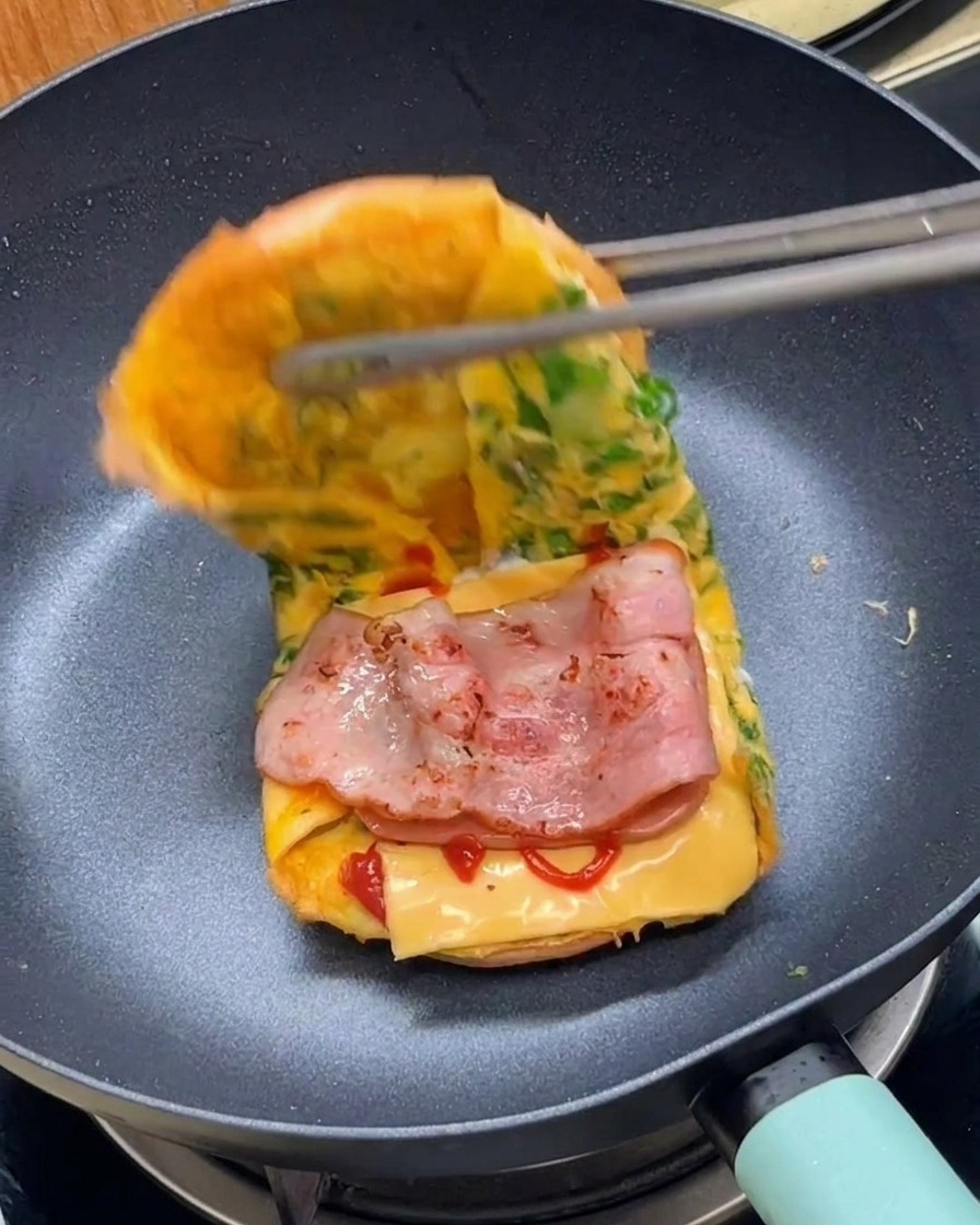 Eine Zange klappt das Ei-Brötchen-Sandwich in der Pfanne zusammen.