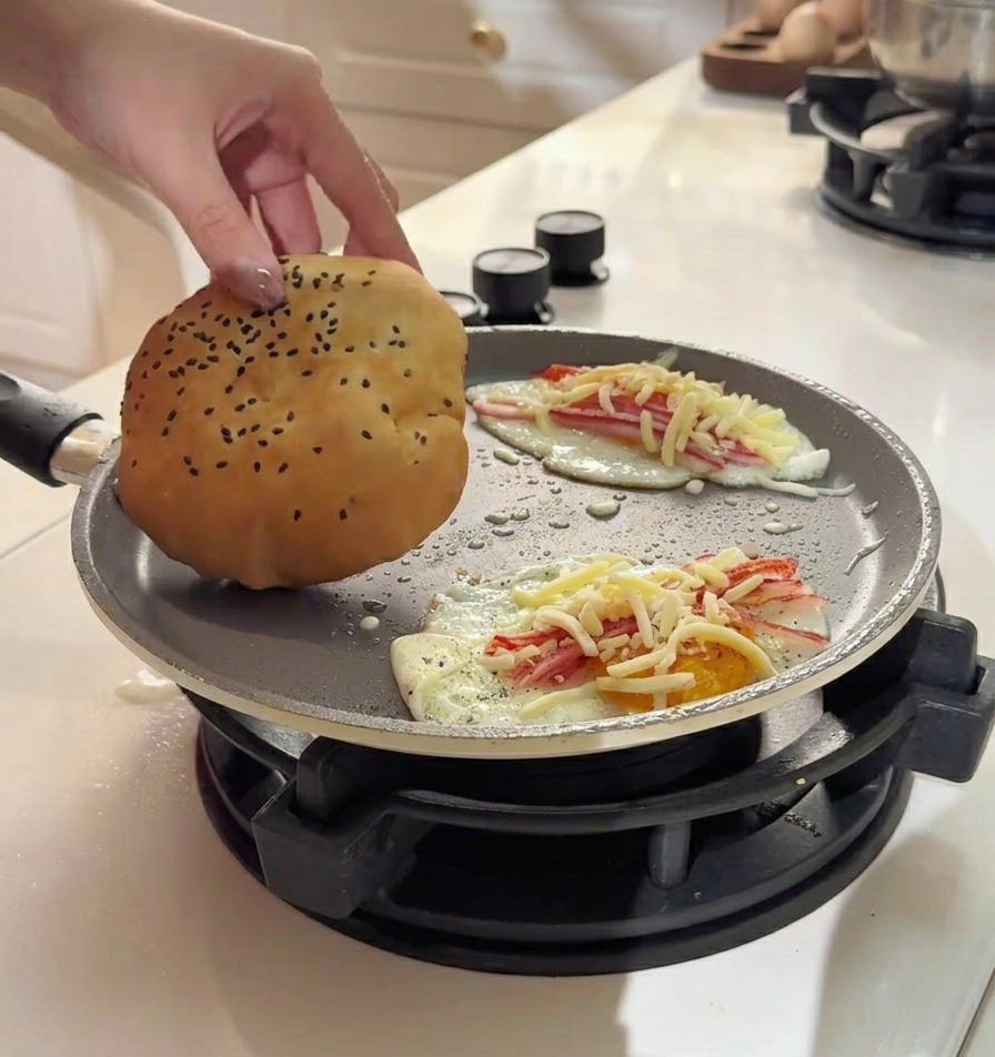 Eine Hand legt ein weiches, goldbraunes Brötchen direkt auf ein Spiegelei mit Belag in der Pfanne.