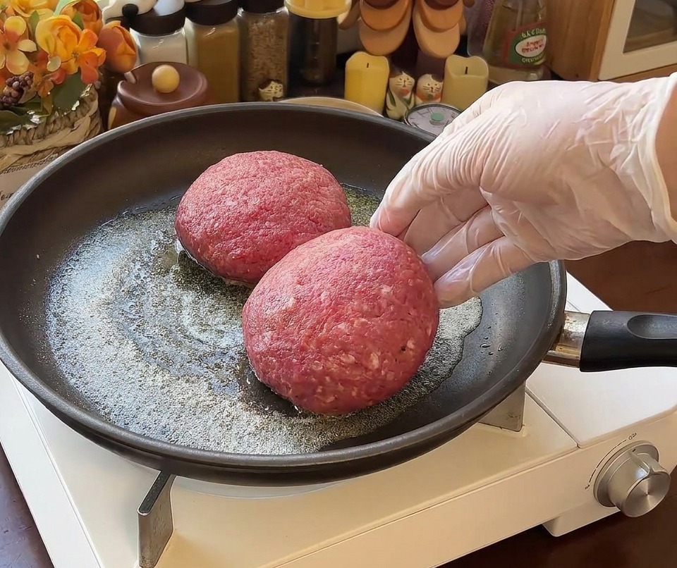 Zwei dicke Rindfleisch-Patties werden in eine Pfanne mit geschmolzener Butter gelegt, während eine Hand assistiert.