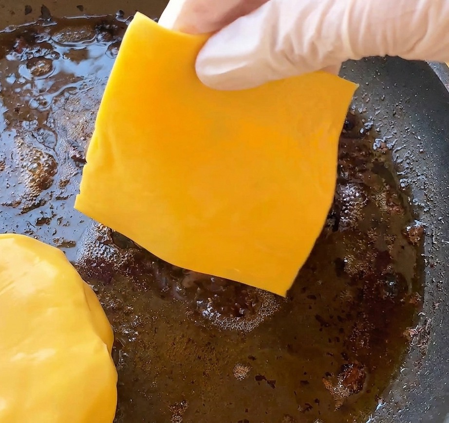 Eine Hand legt eine Scheibe gelben Cheddar-Käse auf ein brutzelndes Burger-Patty in der Pfanne.
