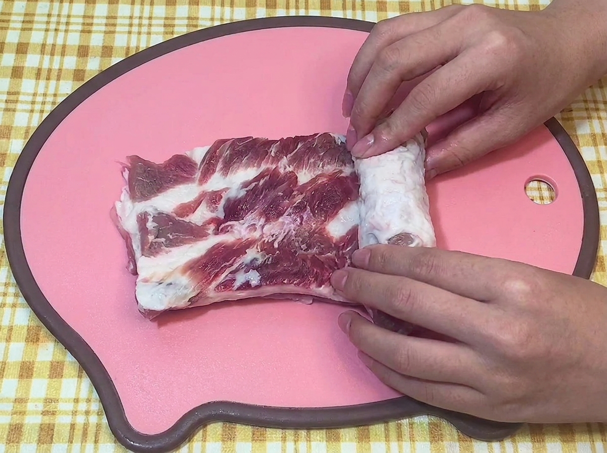 Hände rollen ein rohes Stück Schweinebauch fest zu einem Zylinder auf einem rosa Schneidebrett.