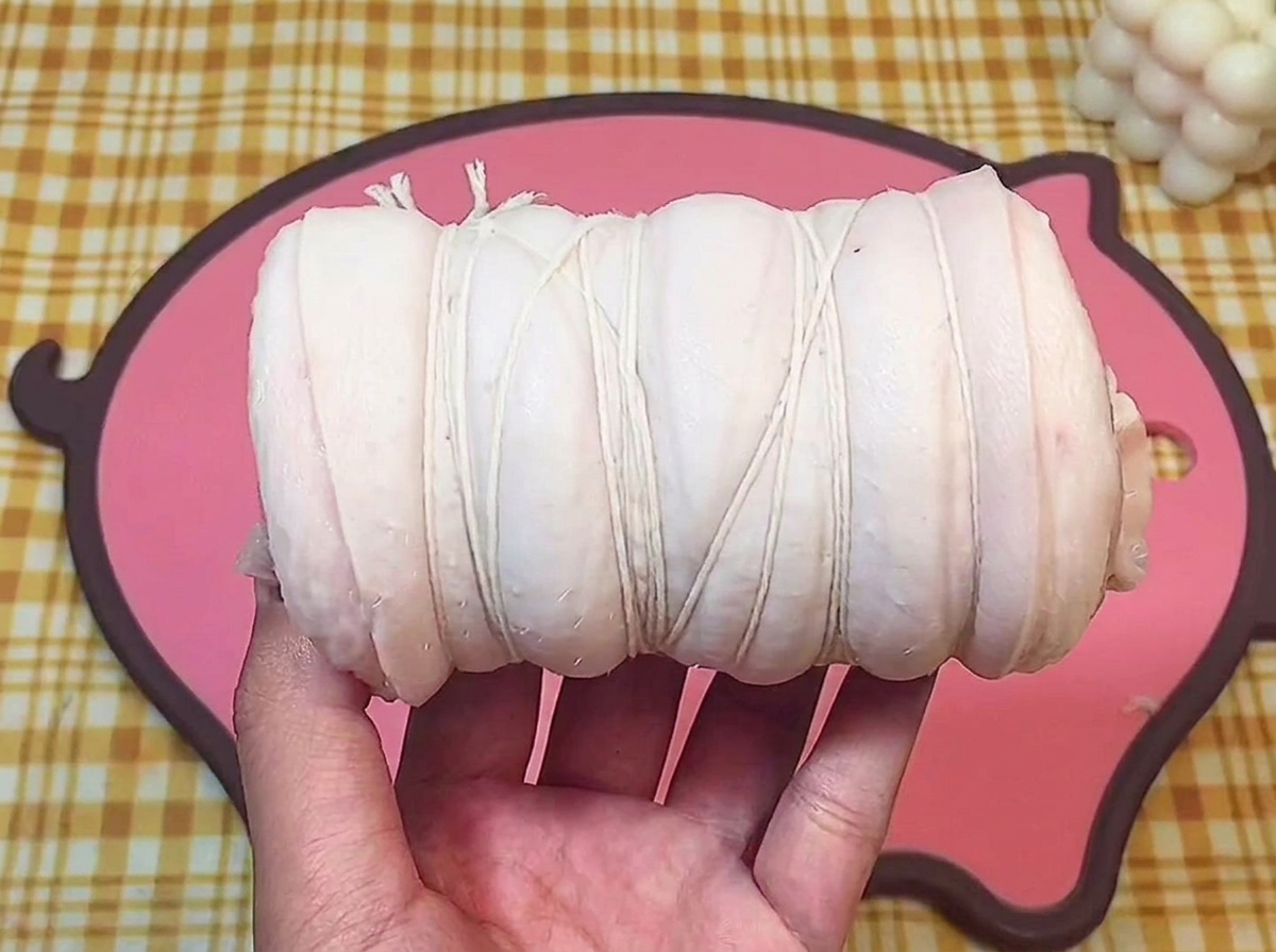 Eine Hand hält den fest gerollten Schweinebauch, der mit weißem Küchengarn fixiert ist.