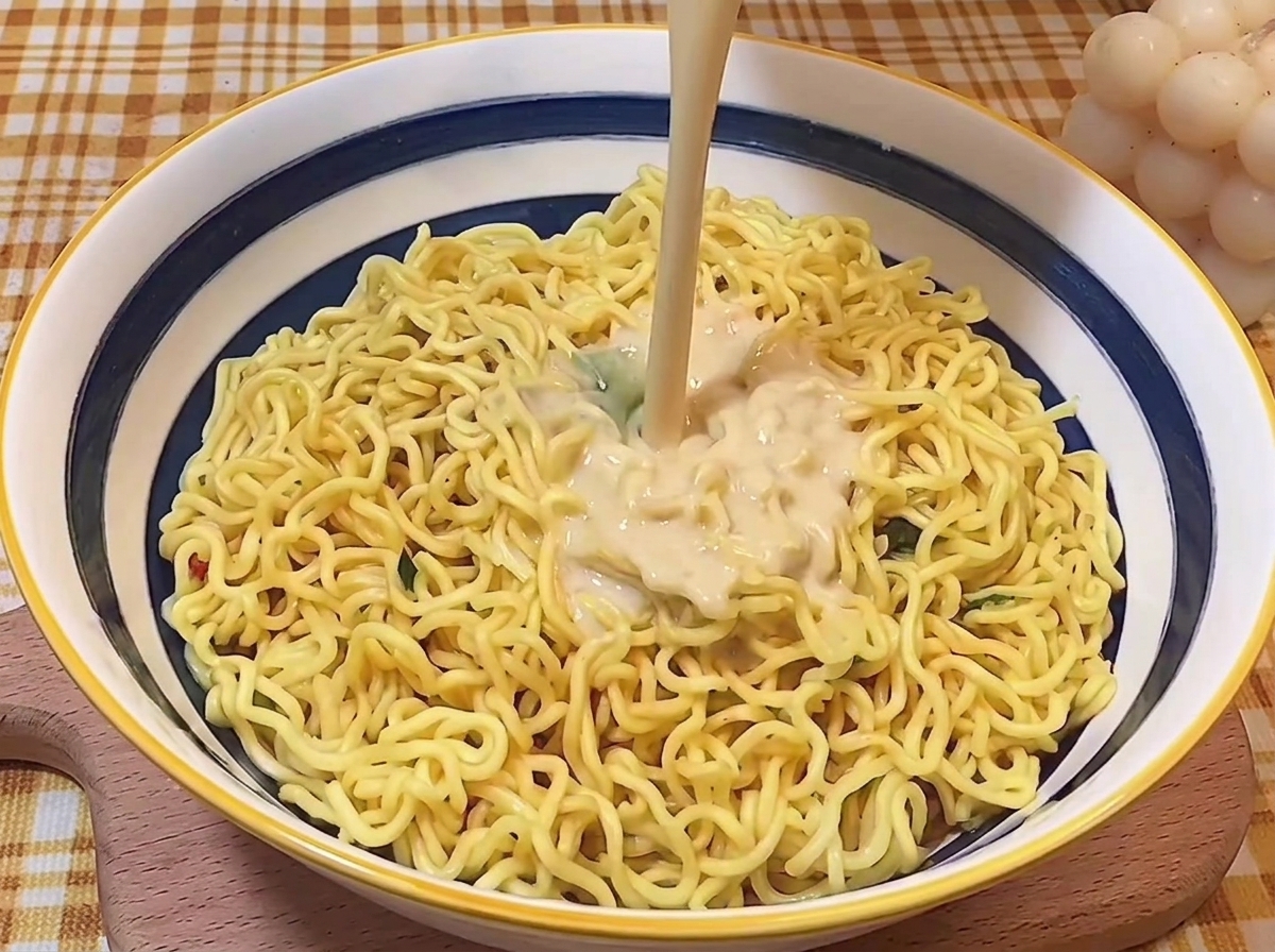 Ein Strahl cremiger Tonkotsu-Brühe wird über die gekochten Nudeln in der Schüssel gegossen.