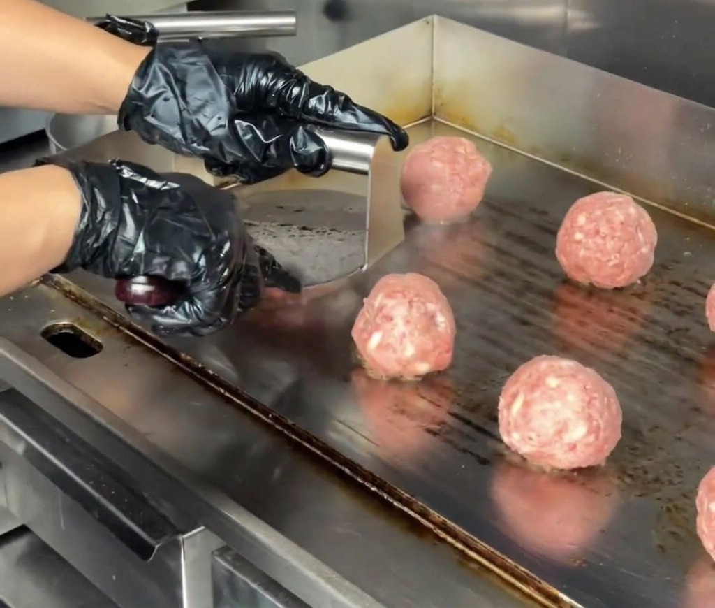 Nahaufnahme einer Burgerpresse aus Edelstahl, die eine Fleischkugel auf einer Grillplatte zu einem dicken Patty flach drückt.