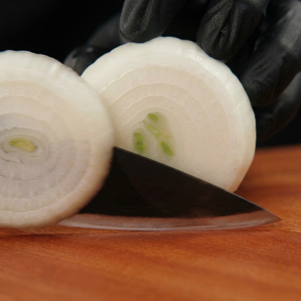 Hände in schwarzen Handschuhen schneiden eine geschälte weiße Zwiebel auf einem Holzbrett in Ringe.