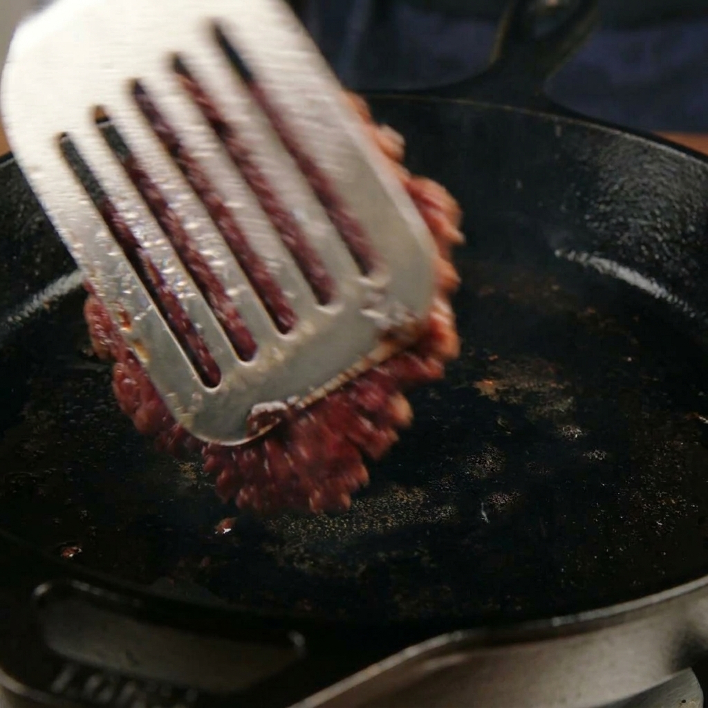 Ein karamellisiertes, flachgedrücktes Rindfleisch-Patty wird mit einem Pfannenwender aus Metall in einer Pfanne gewendet.