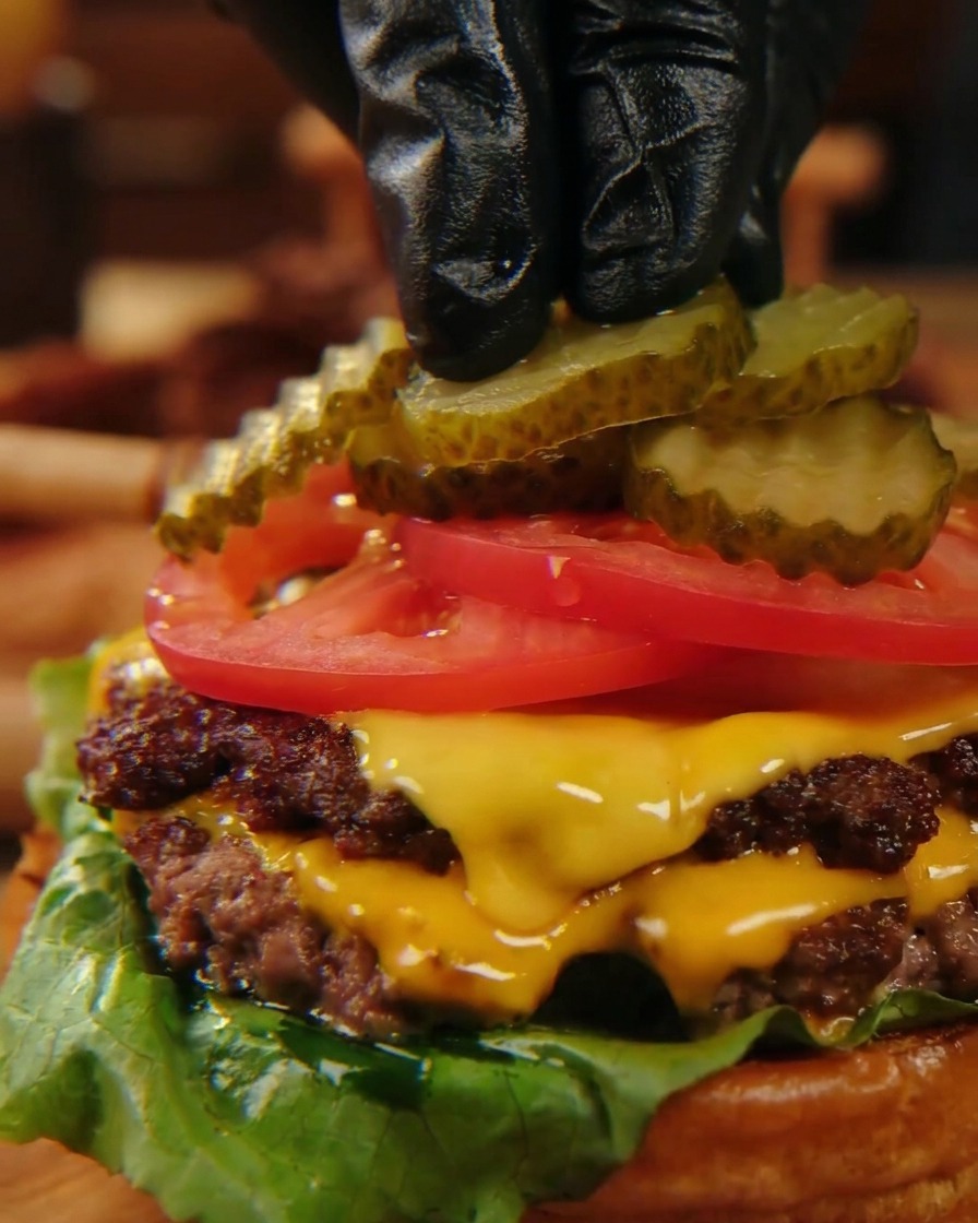 Finger in schwarzen Handschuhen legen vorsichtig geriffelte Gewürzgurken-Scheiben auf leuchtend rote Tomatenscheiben eines geschichteten Cheeseburgers.