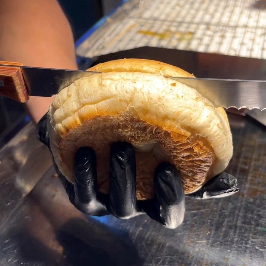 Eine Person schneidet ein weiches Burgerbrötchen mit einem Brotmesser auf einer Edelstahloberfläche horizontal durch.